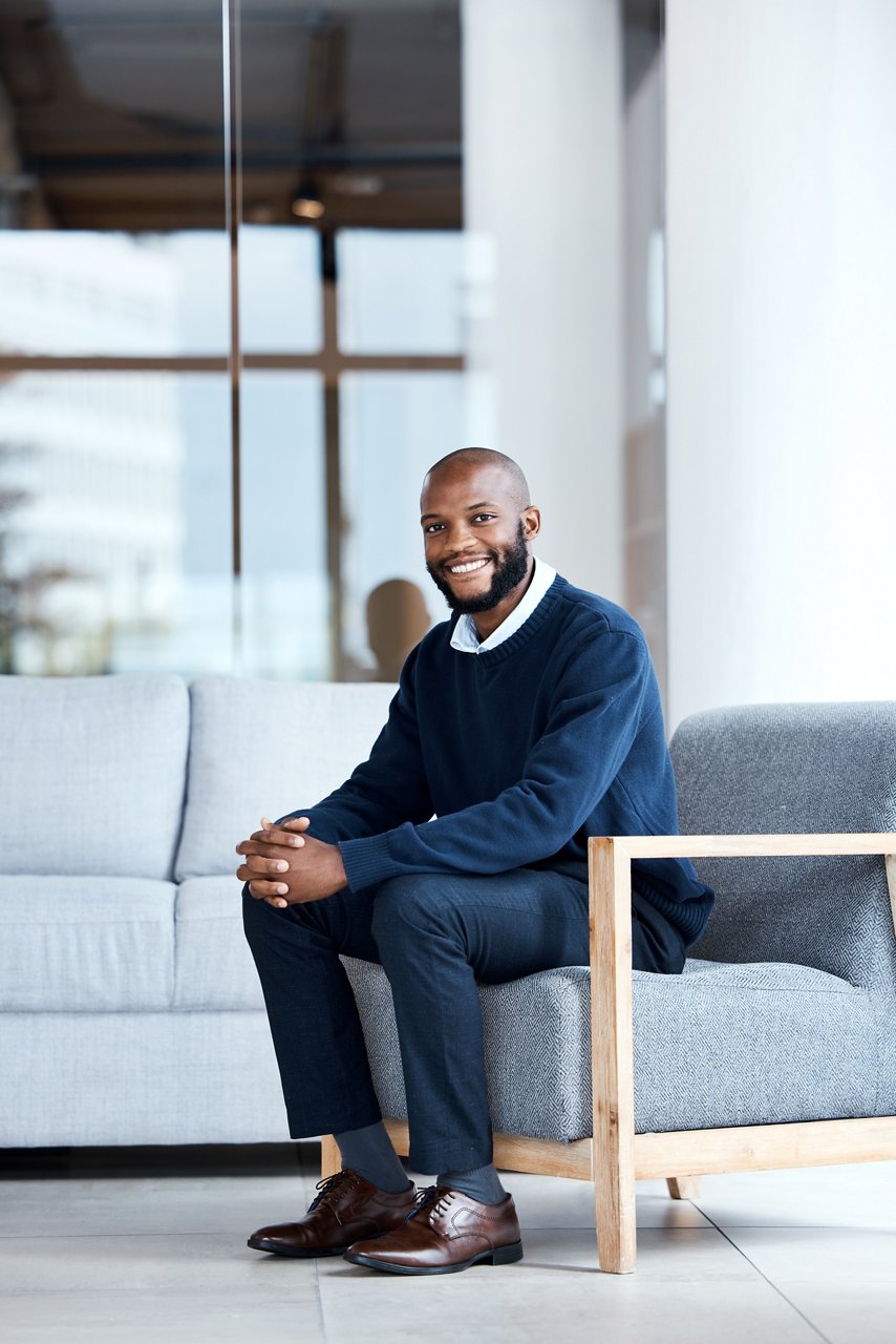 Businessman, happy and smiling at work, job or modern workplace sitting in waiting room for interview at office building. Portrait, employee and African American worker with positive mindset