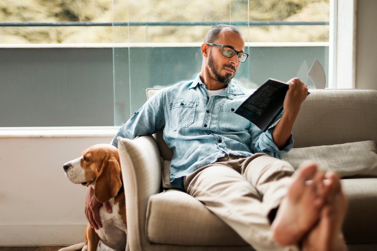 Bearded man comfortably sitting on a coach reading a book and holding his dog .