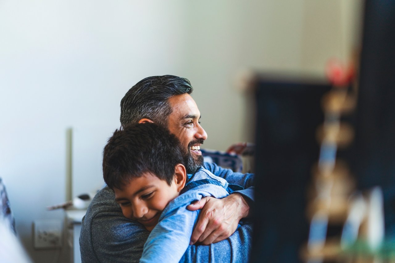 Smiling man embracing boy in bedroom. Happy father is looking away with son at home. They are spending leisure time