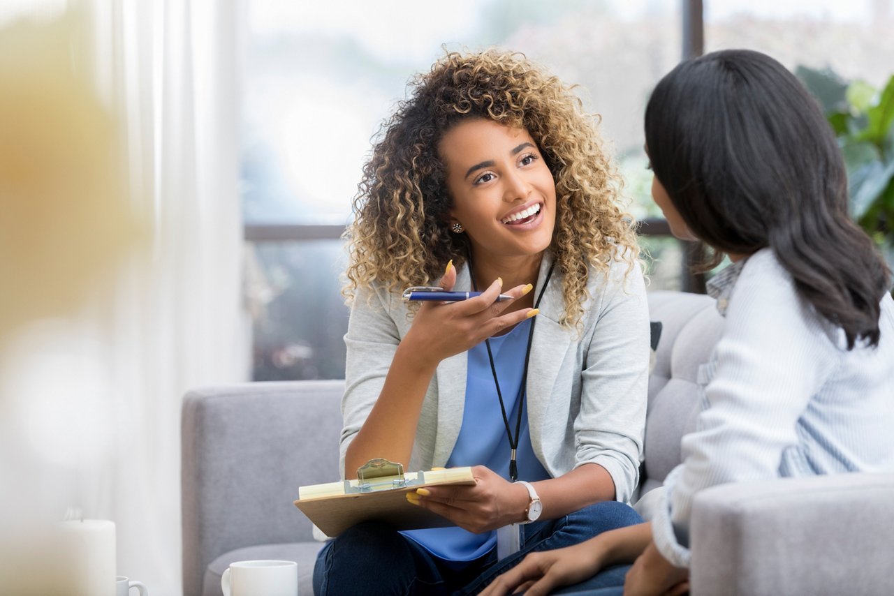 Positive young female therapist gestures as she talks with a female client. The therapist smiles warmly as she talks with the young woman