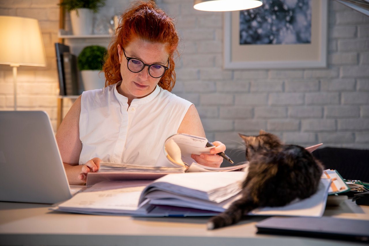 Exhausted, stressed, sleepy woman feeling tired behind her office desk. Working from home. Stressed businesswoman with glasses sitting behind home office desk, cat is watching her