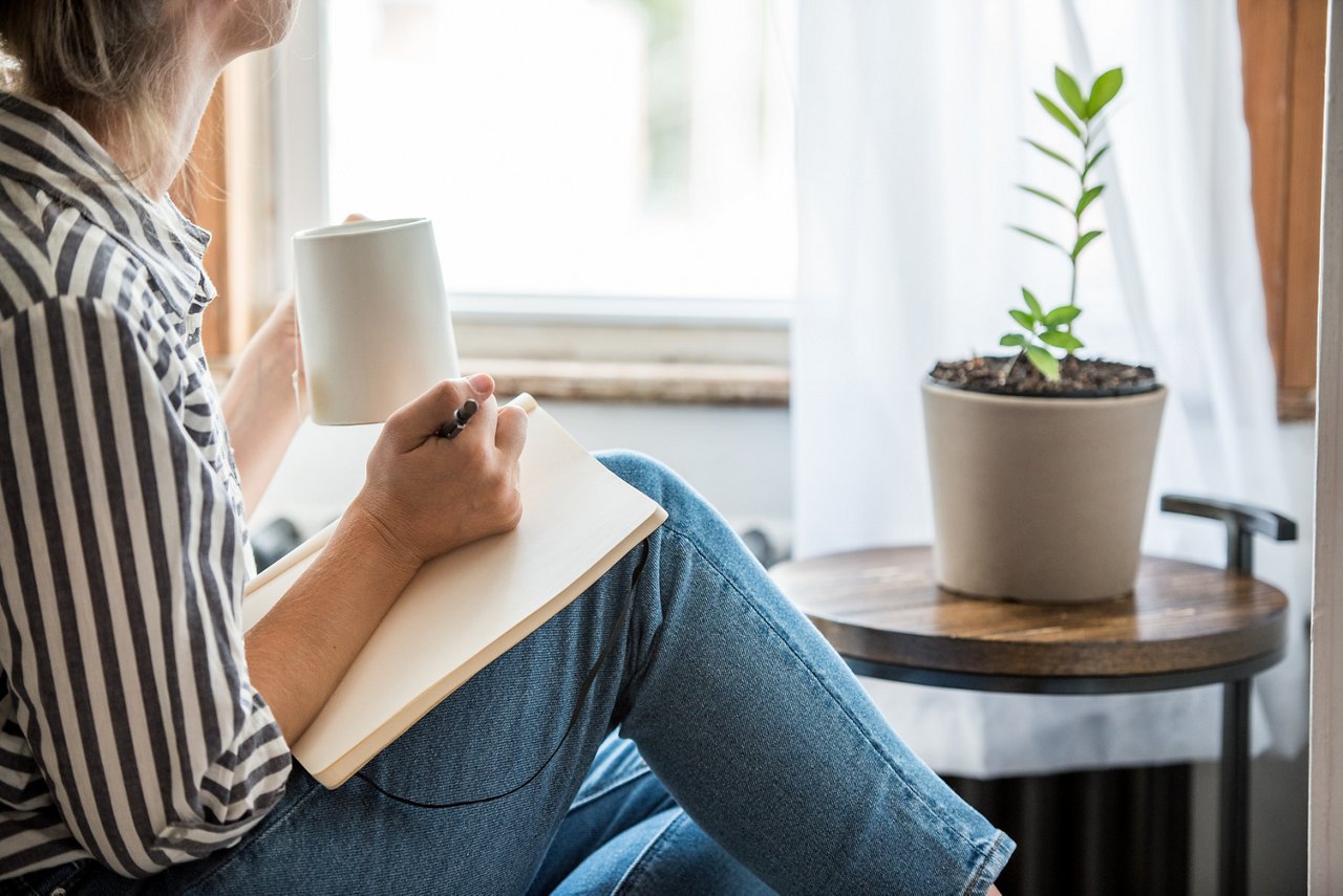 A young woman takes a break to do something analog like writing in her journal and drinking tea. This is a healthy practice for those who experience anxiety